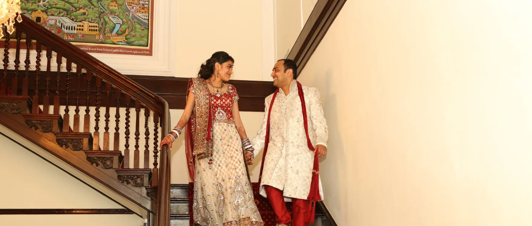 Bride in a red and white lehenga and groom in a cream sherwani on a wooden staircase