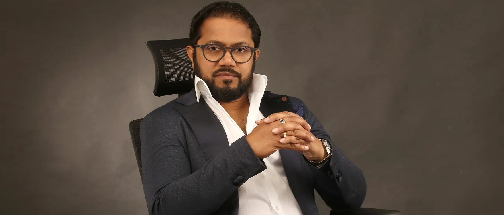 Man in a dark blazer and glasses seated with hands clasped, studio portrait